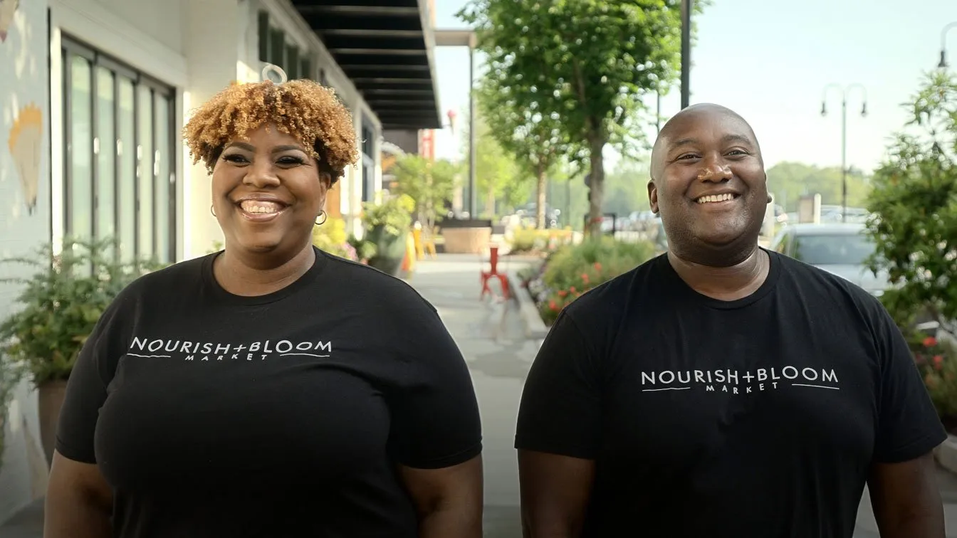 A smiling woman with curly hair and a smiling bald man wearing matching black Nourish + Bloom Market t-shirts stand together outdoors.