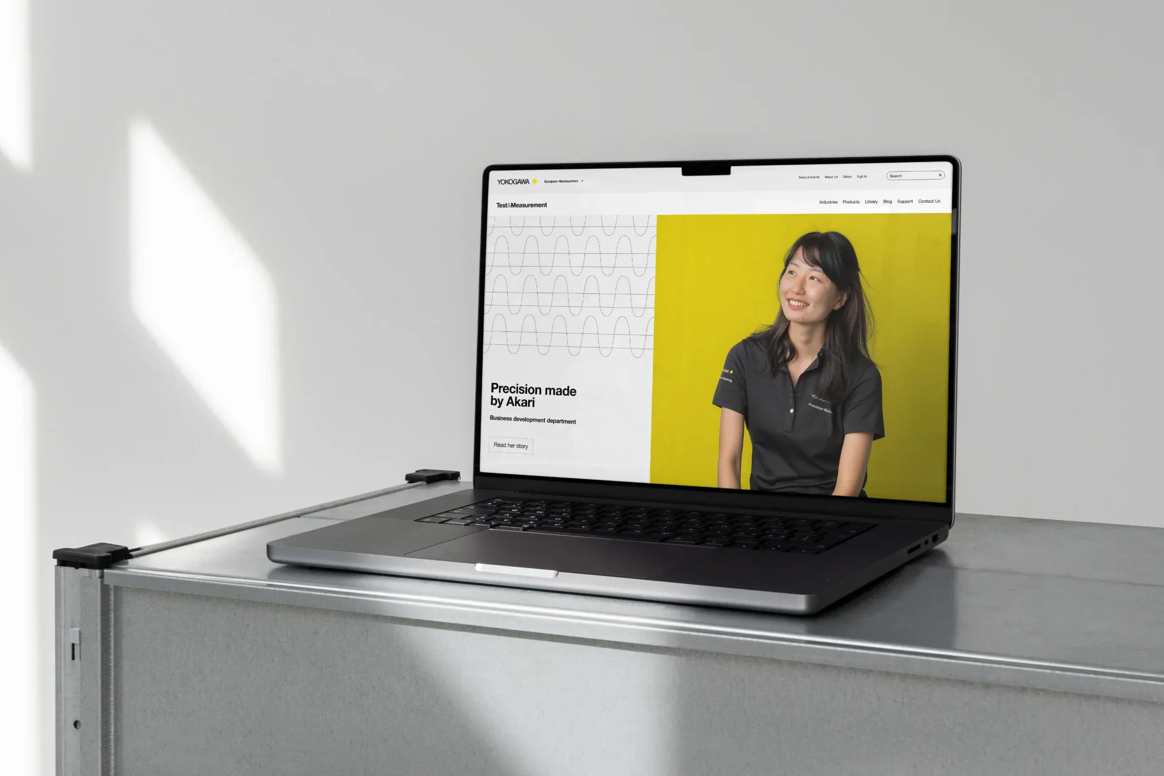A laptop on a metal cabinet showing the Yokogawa Test & Measurement website, with the headline Precision made by Akari beside a portrait on yellow
